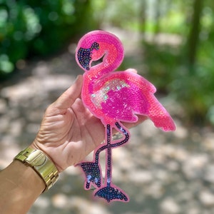 May include: Pink flamingo patch with iridescent sequins. The flamingo is standing on one leg with its head bent down. The patch is made of fabric and has a black outline.