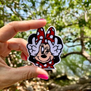 May include: A Minnie Mouse patch with a red and white polka dot bow and dress. Minnie is winking and making a peace sign with her hands. The patch is black, red, white, and tan.