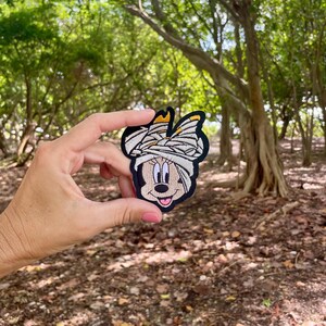 May include: A brown and black embroidered patch of a cartoon character with a white bandage on its head. The character has large black eyes, a pink nose, and a wide smile.