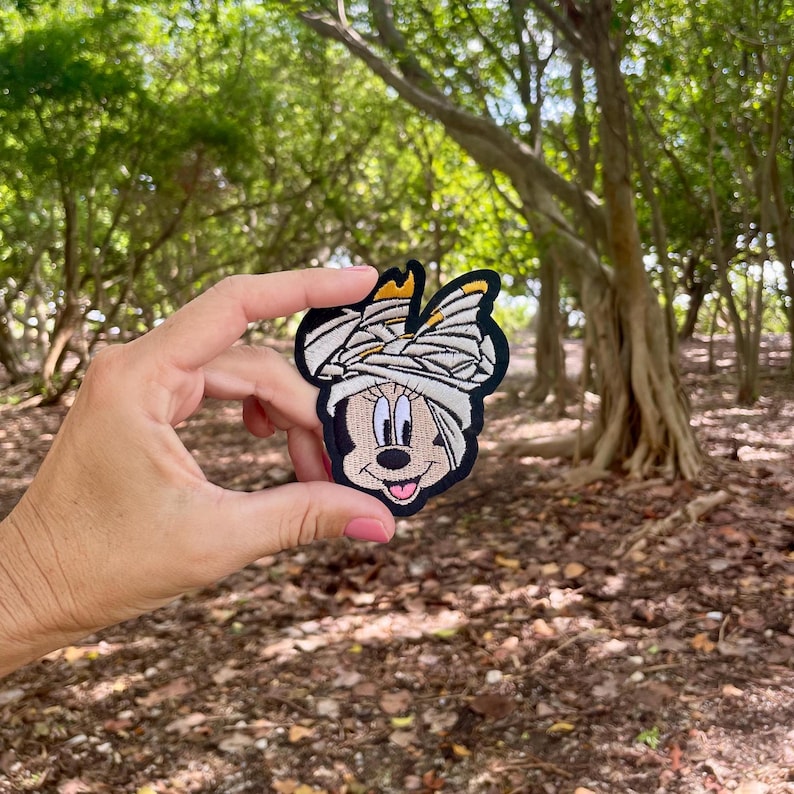 May include: A brown and black embroidered patch of a cartoon character with a white bandage on its head. The character has large black eyes, a pink nose, and a wide smile.