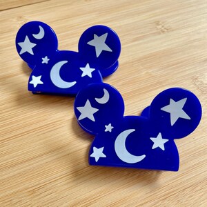 May include: Two blue plastic hair clips shaped like Mickey Mouse ears with white stars and crescent moons on them.