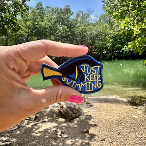 May include: A blue, black, and yellow embroidered patch in the shape of a fish. The patch has the words "Just Keep Swimming" in yellow and blue. The patch is held up in front of a lake.