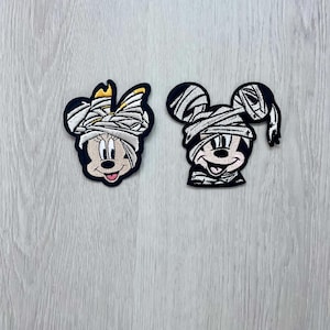 May include: Two embroidered iron-on patches featuring Mickey Mouse and Minnie Mouse dressed as mummies for Halloween. The patches are white and black with a white background and black outlines.