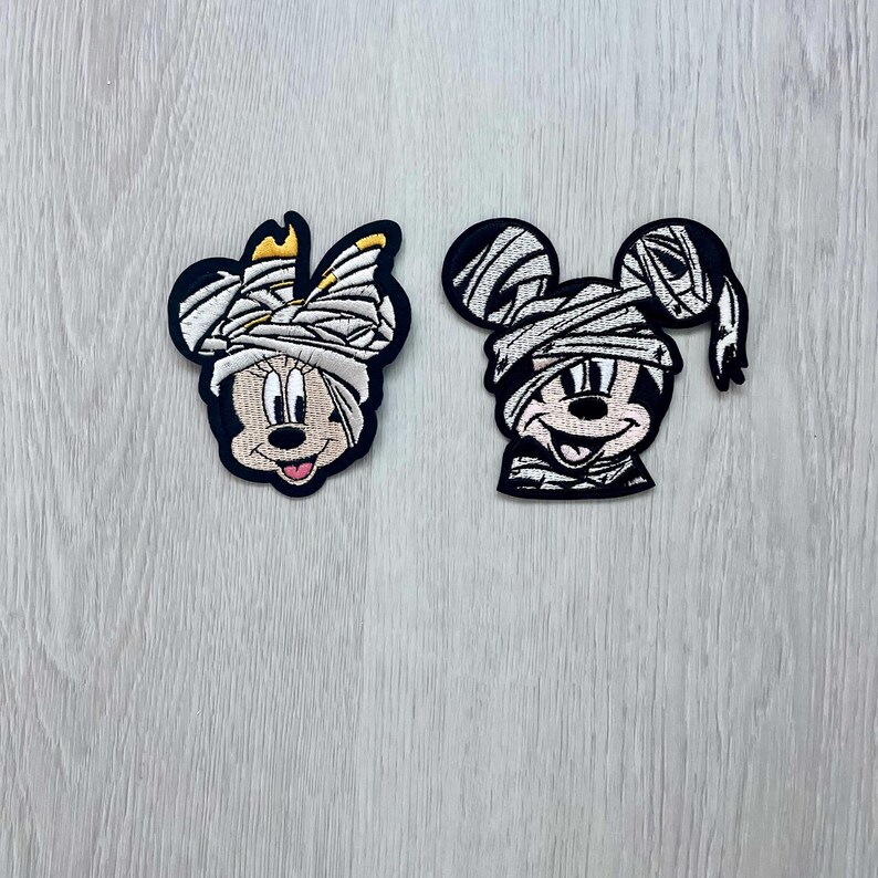 May include: Two embroidered iron-on patches featuring Mickey Mouse and Minnie Mouse dressed as mummies for Halloween. The patches are white and black with a white background and black outlines.