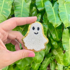 May include: A white ghost with a gold outline and a black smiling face. The ghost is made of fluffy material and is a cute Halloween decoration.