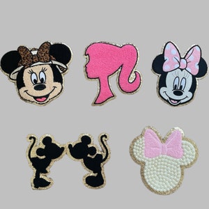 May include: Five iron-on patches featuring cartoon characters. The patches are in various colors, including pink, black, white, and brown. The characters include a mouse wearing a safari hat, a mouse with a pink bow, a mouse with a pink bow and pearls, a silhouette of two mice kissing, and a pink silhouette of a woman with long hair.
