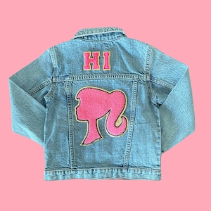 May include: A light blue denim jacket featuring a pink chenille patch of a silhouette profile on the back. Above the silhouette, the word HI is embroidered in pink block letters with a gold border. The jacket has a collar and long sleeves.
