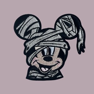 May include: A black and white embroidered patch of a cartoon mouse dressed as a mummy. The mouse is smiling with its tongue sticking out. The patch is perfect for adding a spooky touch to any project.
