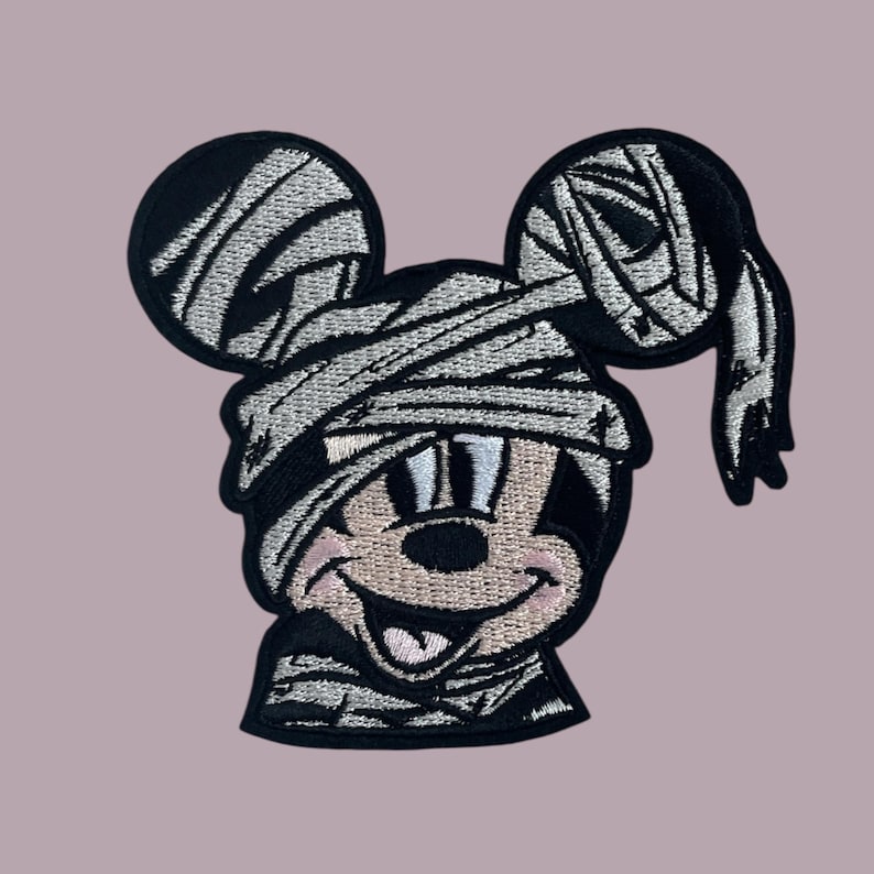 May include: A black and white embroidered patch of a cartoon mouse dressed as a mummy. The mouse is smiling with its tongue sticking out. The patch is perfect for adding a spooky touch to any project.
