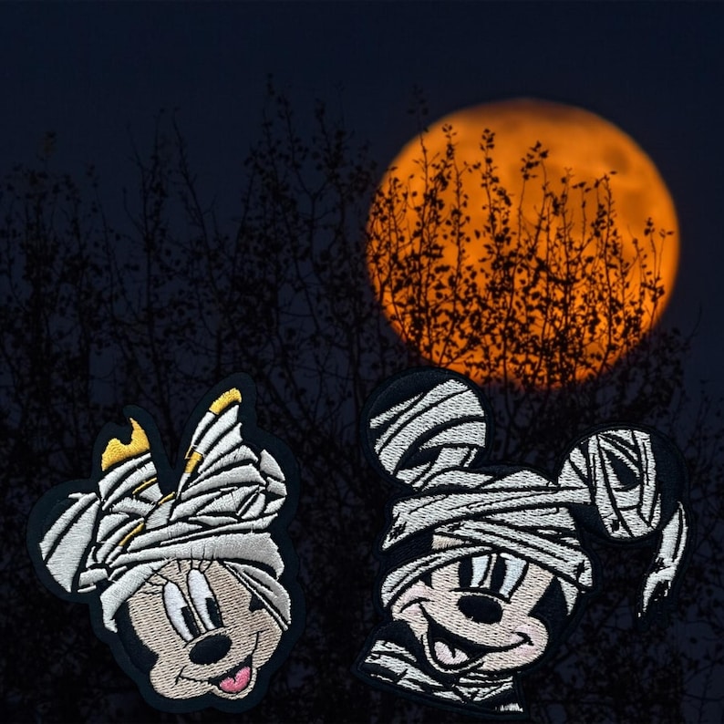 May include: Two embroidered patches of Mickey Mouse and Minnie Mouse dressed as mummies. The patches are white with black outlines and are set against a dark background with a large orange moon.