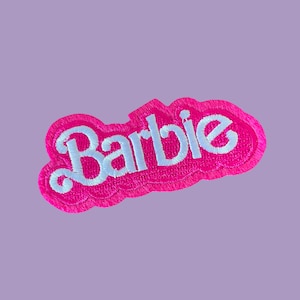 May include: A pink embroidered patch with the word "Barbie" in white script. The patch has a cloud-like shape and is set against a light purple background. The Barbie logo is a classic design.