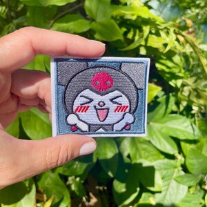 K&ouml;nnte beinhalten: Gestickter B&uuml;gel-Patch mit einer Cartoon-Figur mit einem Sch&auml;del auf dem Kopf, die l&auml;chelt, die Augen geschlossen hat und rote Wangen hat. Die Figur ist von einem blauen und wei&szlig;en Rand umgeben.
