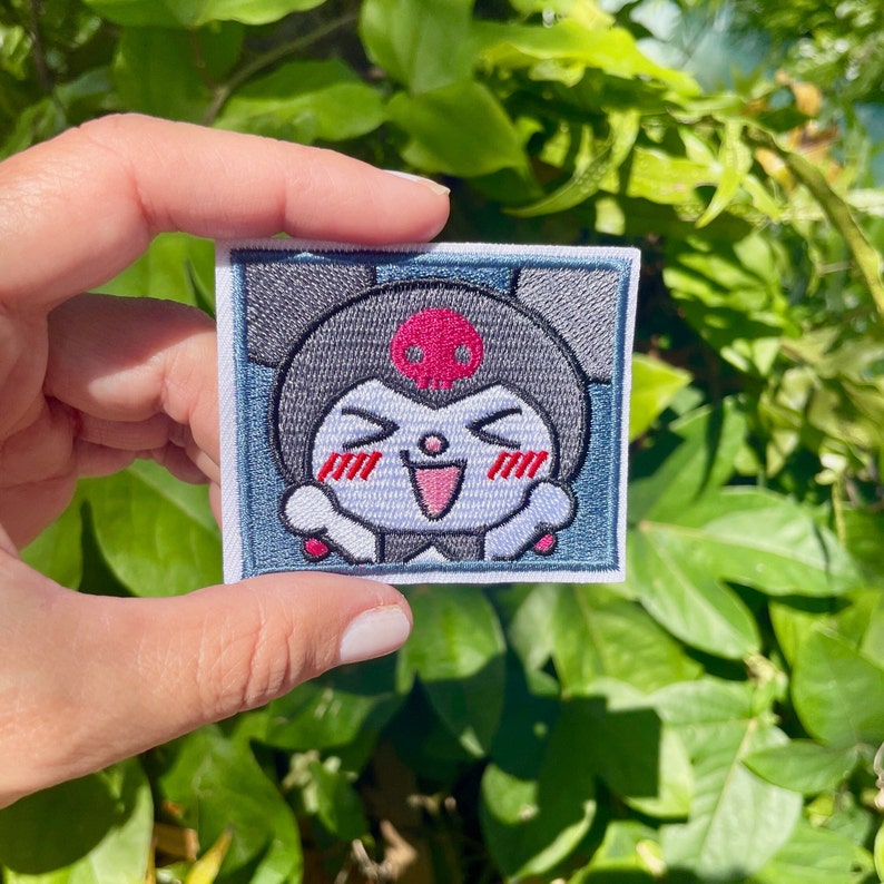 K&ouml;nnte beinhalten: Gestickter B&uuml;gel-Patch mit einer Cartoon-Figur mit einem Sch&auml;del auf dem Kopf, die l&auml;chelt, die Augen geschlossen hat und rote Wangen hat. Die Figur ist von einem blauen und wei&szlig;en Rand umgeben.