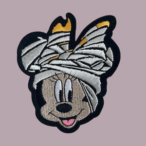 May include: A black and white embroidered patch of a cartoon mouse with a white bandage wrapped around its head. The bandage is decorated with a gold bow.