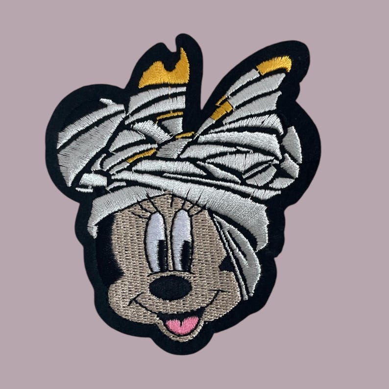 May include: A black and white embroidered patch of a cartoon mouse with a white bandage wrapped around its head. The bandage is decorated with a gold bow.