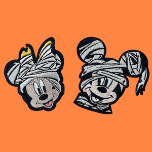 May include: Two embroidered iron-on patches featuring Mickey Mouse and Minnie Mouse dressed as mummies for Halloween. The patches are black with white and gray details.