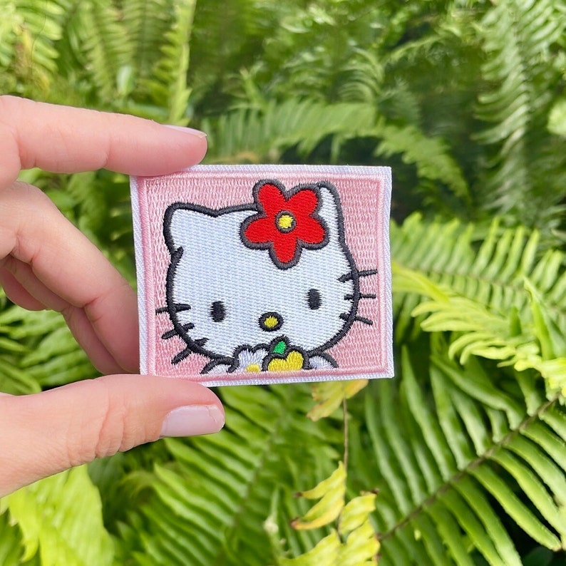 K&ouml;nnte beinhalten: Ein rosa-wei&szlig; gestickter B&uuml;gel-Patch mit Hello Kitty, die eine gelbe Blume mit einer roten Blume auf dem Kopf h&auml;lt.