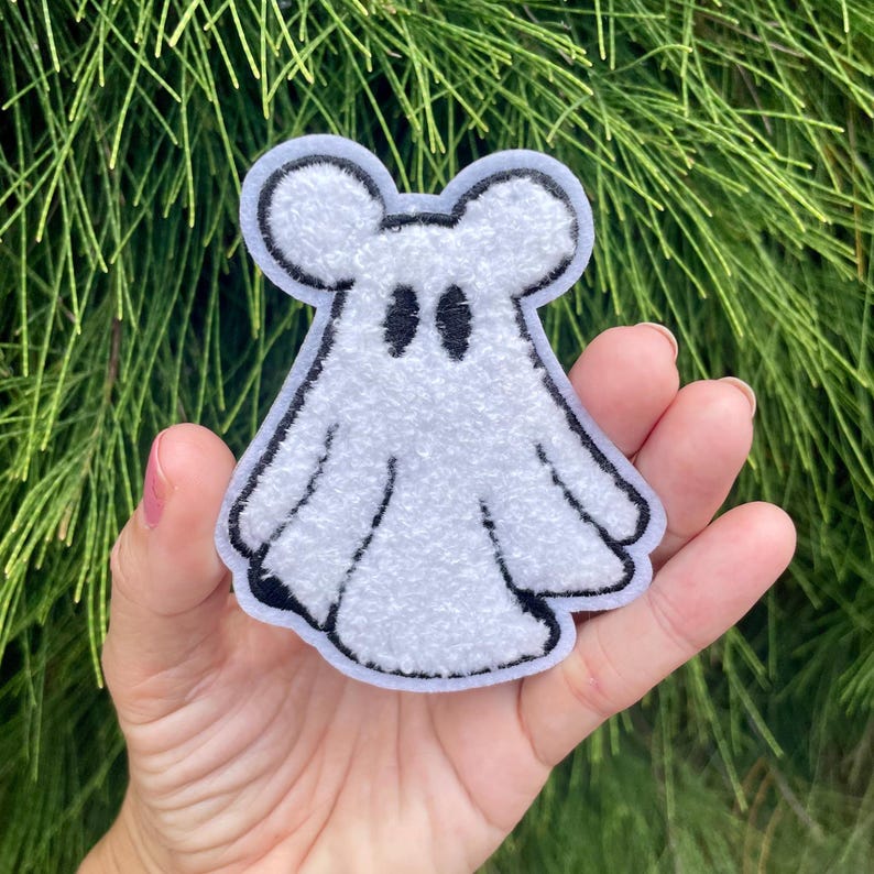 May include: A white fuzzy ghost with black eyes and a black outline. The ghost has two small ears on top of its head. The ghost is a patch or iron-on.
