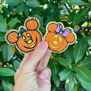 May include: Two felt pumpkin patches with Mickey Mouse ears. One pumpkin is orange with a purple bow and the other is orange with a green stem.