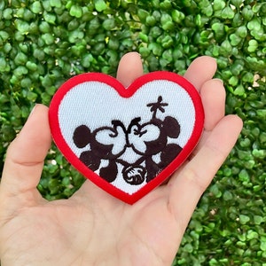 May include: Red heart-shaped embroidered patch with a white background and a black silhouette of two cartoon characters kissing. The characters are holding hands and there is a small palm tree above them.