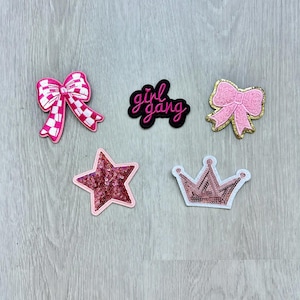 May include: Five iron-on patches with pink and white designs. The patches include a bow, a star, a crown, a bow with gold accents, and a black patch with the text "girl gang" in pink.