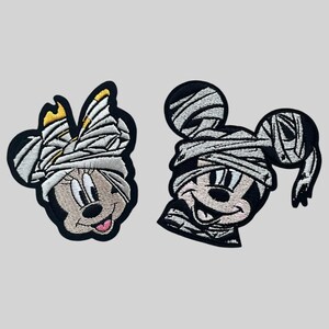May include: Two embroidered iron-on patches featuring Mickey Mouse and Minnie Mouse dressed as mummies. The patches are black and white with a cartoon style.