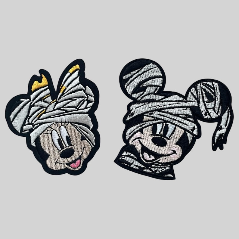 May include: Two embroidered iron-on patches featuring Mickey Mouse and Minnie Mouse dressed as mummies. The patches are black and white with a cartoon style.