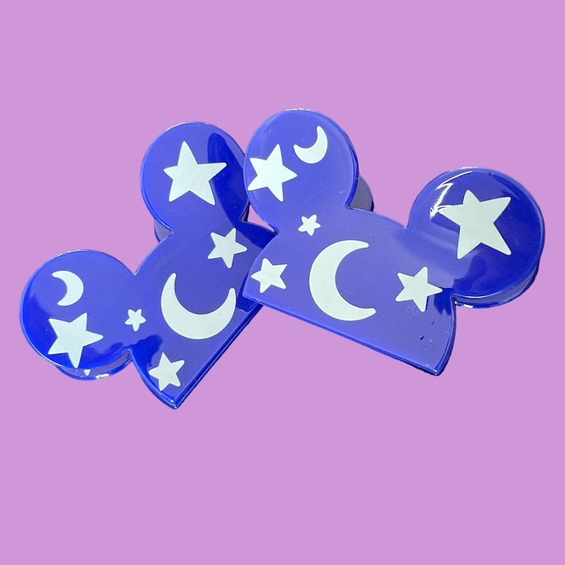 May include: Two blue plastic hair clips shaped like Mickey Mouse ears. The clips are decorated with white stars and crescent moons.
