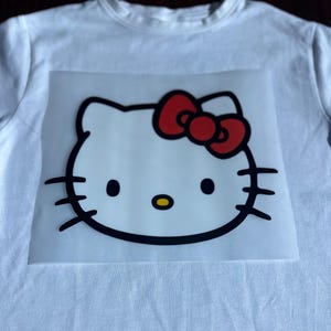 K&ouml;nnte beinhalten: Wei&szlig;es T-Shirt mit einem transparenten B&uuml;gelbild von Hello Kitty mit einer roten Schleife.