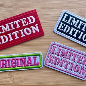 May include: Four embroidered patches with white stitching on a wood background. The patches are rectangular and have different colors and text. One patch is red with white text that says "LIMITED EDITION". Another patch is black with white text that says "LIMITED EDITION". A third patch is white with pink text that says "LIMITED EDITION". The last patch is white with pink text that says "ORIGINAL" and a green border.