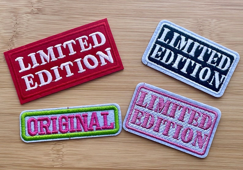 May include: Four embroidered patches with white stitching on a wood background. The patches are rectangular and have different colors and text. One patch is red with white text that says "LIMITED EDITION". Another patch is black with white text that says "LIMITED EDITION". A third patch is white with pink text that says "LIMITED EDITION". The last patch is white with pink text that says "ORIGINAL" and a green border.