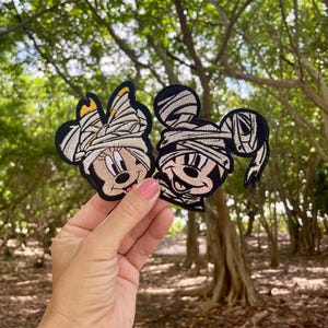 May include: Two embroidered patches of Mickey Mouse and Minnie Mouse dressed as mummies. The patches are beige and black with white stitching.