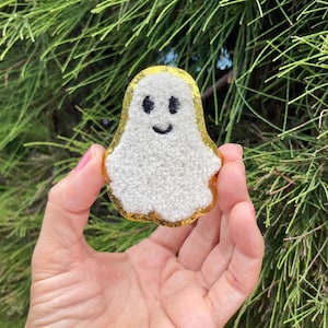 May include: A white fuzzy ghost with a gold glitter outline and a smiling face. The ghost is held in a hand.
