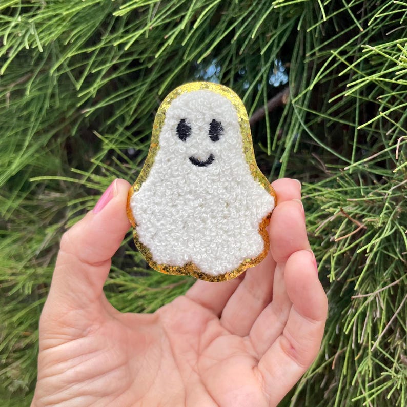 May include: A white fuzzy ghost with a gold glitter outline and a smiling face. The ghost is held in a hand.