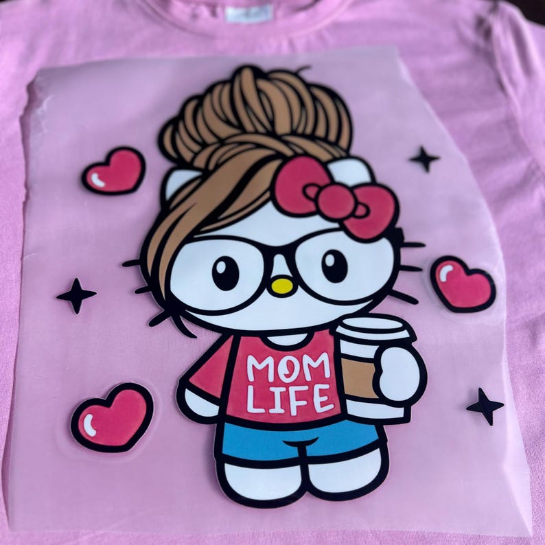 K&ouml;nnte beinhalten: Ein wei&szlig;er Hello Kitty Charakter mit braunen Haaren in einem unordentlichen Dutt, der eine rosa Brille und ein rotes Shirt mit dem Text "MOM LIFE" tr&auml;gt und eine braune Kaffeetasse h&auml;lt. Der Charakter ist von rosa Herzen und Sternen umgeben.