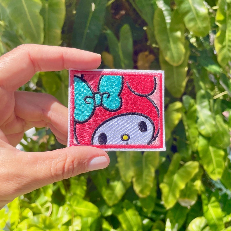 K&ouml;nnte beinhalten: Ein gestickter Aufn&auml;her mit einem Comic-Charakter mit einer blauen Schleife. Der Charakter ist rot und wei&szlig; mit gro&szlig;en Augen und einer kleinen gelben Blume auf der Nase.