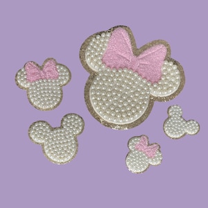 May include: Five pearl and glitter iron-on patches in the shape of a cartoon mouse head with a pink bow. The patches are perfect for adding a touch of whimsy to clothing, bags, or other accessories.