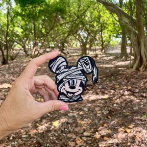 May include: A black and white embroidered patch of a cartoon character dressed as a mummy. The character has large ears and a wide smile. The patch is held in a hand against a background of trees.