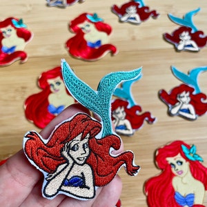May include: A set of nine embroidered iron-on patches featuring Ariel, the Little Mermaid. The patches are in various poses and feature red, blue, and green colors.