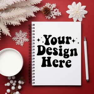 May include: A white spiral-bound notebook with the text "Your Design Here" in black, set against a red background. A white pen, a cup of liquid, and decorative elements like snowflakes and pinecones surround the notebook.