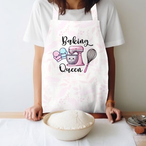 Puede incluir: Delantal blanco con estampado floral rosa y el texto "Baking Queen" en letra negra. Se muestra una batidora de pie rosa con un taz&oacute;n blanco y un batidor. Dos guantes de horno rosa y azul est&aacute;n a un lado de la batidora.