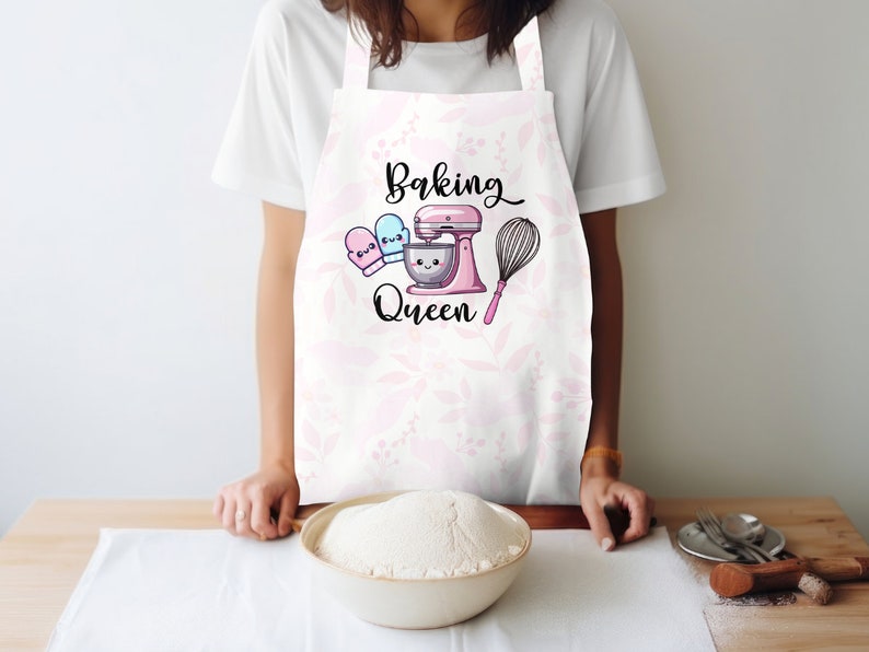 Puede incluir: Delantal blanco con estampado floral rosa y el texto "Baking Queen" en letra negra. Se muestra una batidora de pie rosa con un taz&oacute;n blanco y un batidor. Dos guantes de horno rosa y azul est&aacute;n a un lado de la batidora.