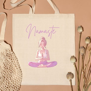 May include: Beige tote bag with the word "Namaste" in lavender script above an illustration of a meditating person. The person is wearing a white top and pink pants. A beige string bag is on the left.