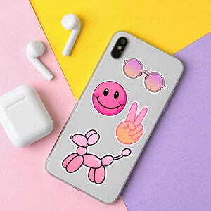Op de afbeelding: Een witte telefoonhoes met roze en oranje stickers. De stickers bevatten een smiley, een vredesteken, een zonnebril en een roze ballondier.