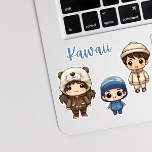 May include: Three cartoon characters wearing winter hats and coats. The characters are a girl wearing a brown coat and a white bear hat, a boy wearing a blue coat and a blue hat, and a boy wearing a white coat and a white hat. The text "Kawaii" is written in blue letters on a white background.