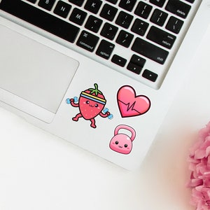 May include: A collection of fitness-themed stickers on a laptop. The stickers include a cartoon strawberry lifting weights, a pink heart with a heartbeat line, and a pink kettlebell. The stickers are colorful and playful.