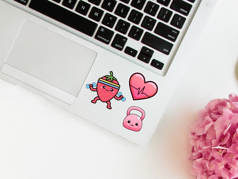 May include: A collection of fitness-themed stickers on a laptop. The stickers include a cartoon strawberry lifting weights, a pink heart with a heartbeat line, and a pink kettlebell. The stickers are colorful and playful.