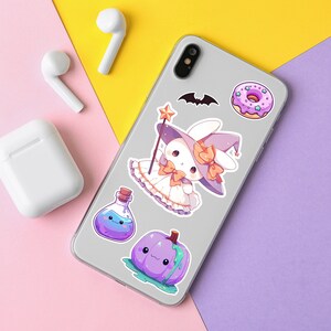 Puede incluir: Una funda de tel&eacute;fono blanca con una variedad de pegatinas de colores. Las pegatinas incluyen una dona morada y blanca, una calabaza morada, una botella de poci&oacute;n morada y un conejito blanco con un sombrero de bruja morado y una varita.