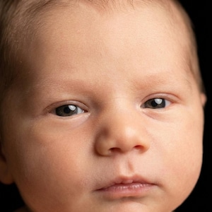 May include: A close-up portrait of a newborn baby with fair skin and dark eyes. The baby's face is centered, with a soft focus on the features. The background is solid black, highlighting the baby's delicate features.