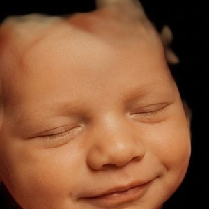 May include: A close-up of a baby's face with closed eyes and a slight smile. The skin is a warm, peachy tone against a black background. The image captures the soft features of a newborn.